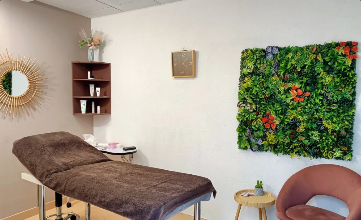 La cabine de Priscillia Beauté : table de soin, miroir soleil doré, étagère de produits et mur végétal fleuri